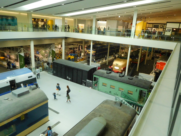 Ein ger├Ąumiger Museumsausstellungsraum mit verschiedenen Zügen, Menschen, Gel├Ąndern, Pfeilern, Beleuchtung und Informationsschildern.