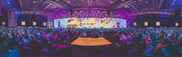Eine große Menschenmenge auf Stühlen sitzend, die einer hell erleuchteten Bühne mit mehreren stehenden Individuen zugewandt sind, wahrscheinlich bei einem Online-Marketing-Rockstars-Event umgeben von Bildschirmen.