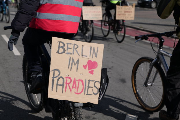 Eine Gruppe von Menschen, die auf Fahrrädern eine Straße entlangfahren, mit einem "Berlin I'm Paradies"-Schild im Vordergrund, einem Auto im Hintergrund und dem Bild etwas unscharf.