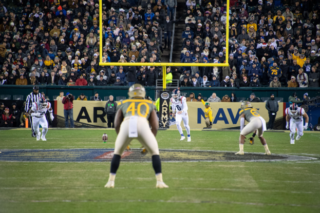 Fußballspieler mit Helmen und Uniformen auf einem Feld mit Zuschauern auf den Tribünen, einschließlich sichtbarer Beschilderung und Stützstrukturen.