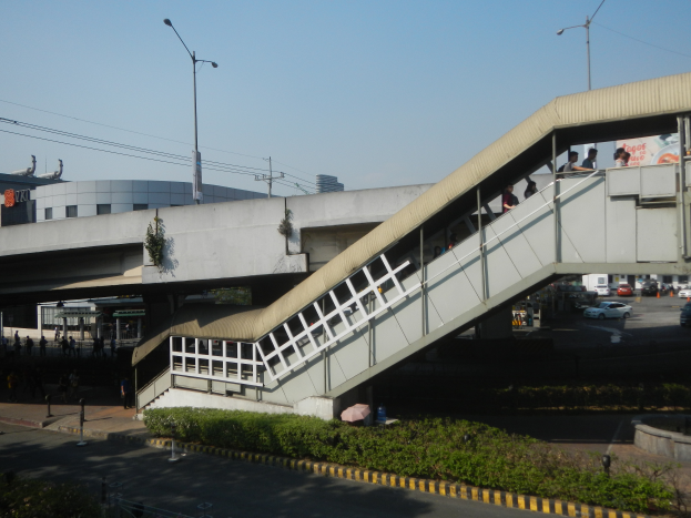 Eine Fußgängerbrücke mit Menschen darauf, die über eine Straße gehen, umgeben von Gebäuden, Straßenlaternen, Strommasten, Stromkabeln, Kraftfahrzeugen, Büschen und einem klaren blauen Himmel.