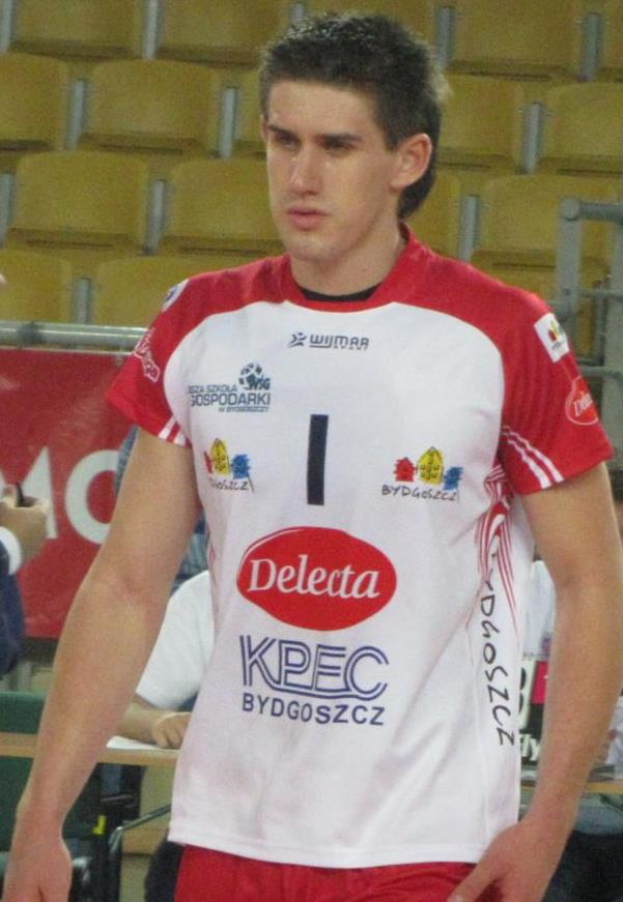 Ein Mann in einer roten und weißen Handballuniform steht auf einem Feld, sieht entschlossen aus, mit Zuschauern und einem roten Banner mit der Aufschrift "Krasnoyarsk vs Bydgoszcz" im Hintergrund.