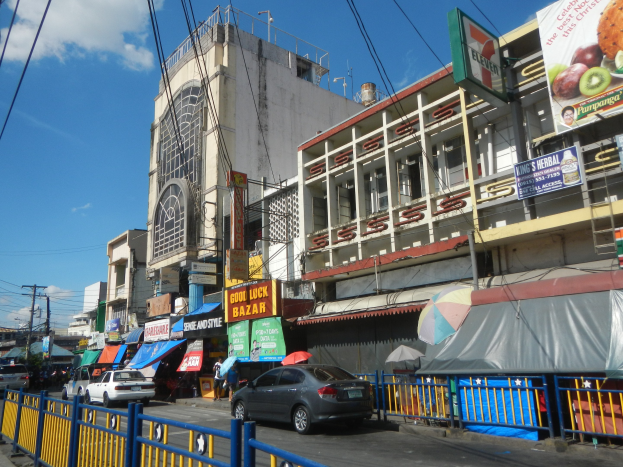 Eine belebte Stadtstraße mit Fahrzeugen, hohen Gebäuden mit Fenstern und Schildern, Fußgängern mit Regenschirmen, einem Metallzaun, Strommasten mit Drähten und einem bewölkten Himmel.