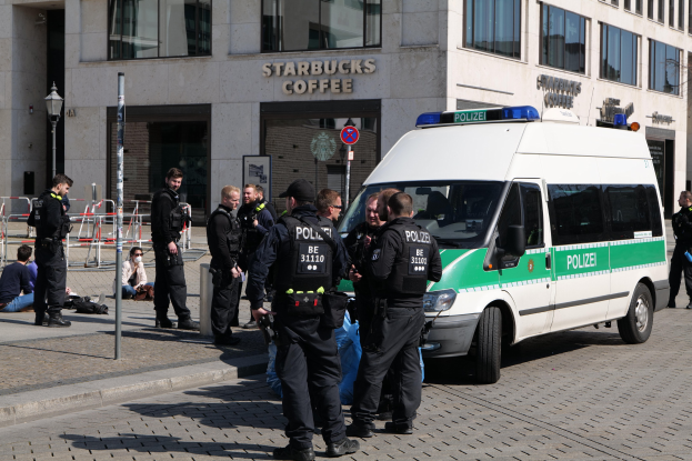Eine Gruppe von Polizisten steht vor einem Starbucks-Café, rechts ein Van und links ein paar Menschen; im Hintergrund ist ein Gebäude mit Fenstern, ein Schild, eine Laterne und ein Zaun zu sehen.