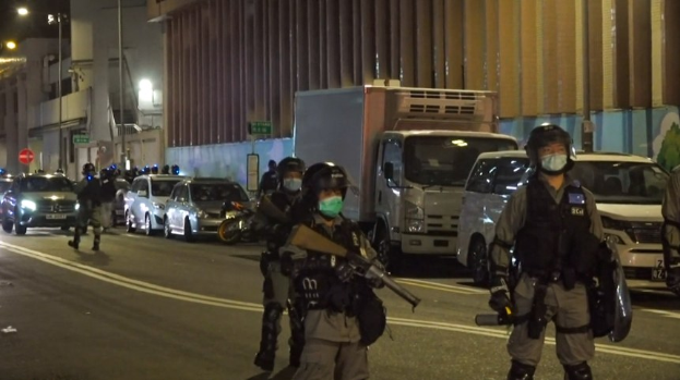 Eine Gruppe von Polizisten in Helmen, Handschuhen und Gesichtsmasken steht auf einer nächtlichen Stadtstraße, einige halten Waffen, mit Fahrzeugen, Laternenmasten, Schildern und Gebäuden im Hintergrund.