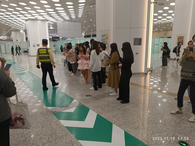Menschen in der Schlange an einem Flughafen-Sicherheitscheck-in, einige mit Taschen und Mobiltelefonen, mit Säulen und Deckenlampen im Hintergrund.