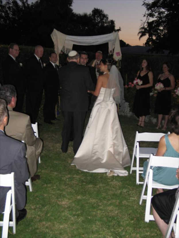 Eine Gruppe von Menschen steht bei einer Hochzeit, wobei die Frauen rechts Blumensträuße halten, während andere auf Stühlen sitzen, umgeben von Bäumen.