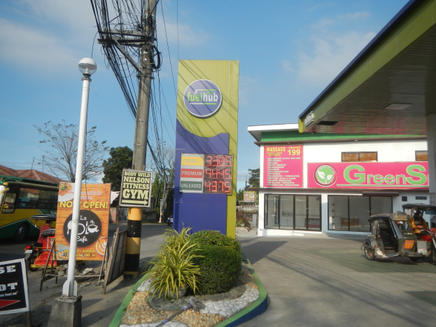 Tankstelle mit einem 'Green's'-Schild, Fahrzeuge auf der Straße, umliegende Infrastruktur und ferne Bäume unter einem bewölkten Himmel.