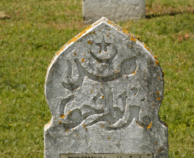 Ein steinerner Grabstein mit arabischer Inschrift, umgeben von Gras auf einem Friedhof.