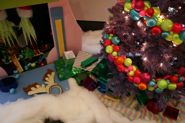 Drinnen, ein geschmückter Weihnachtsbaum rechts, Geschenkboxen und Deko in der Mitte, eine Hütte mit Deko oben links und Baumwolle und Wolle unten.