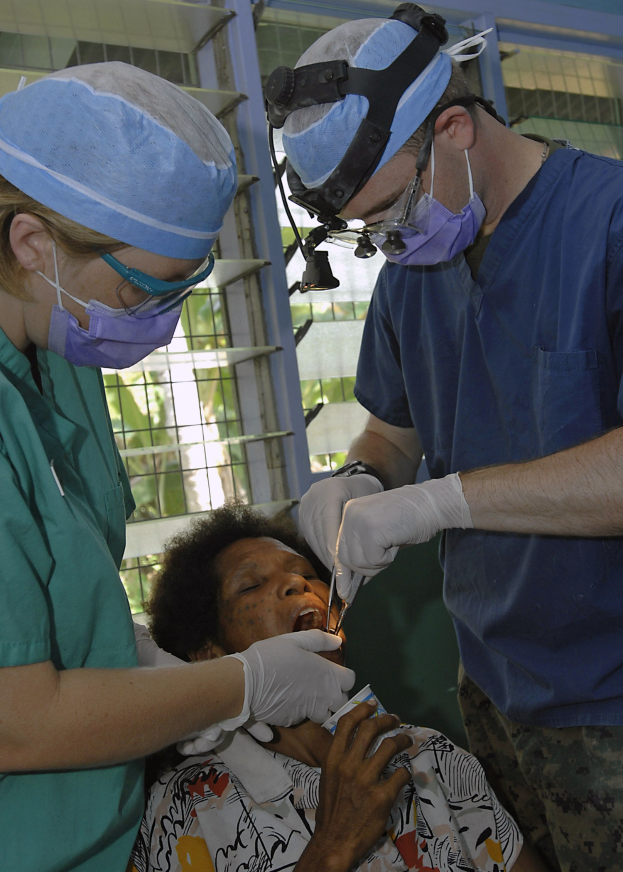 Zwei Chirurgen in Masken und Kappen führen eine Gesichtsoperation an einer Frau durch, die auf einem Bett liegt, mit einem Fenster und Bäumen im Hintergrund.