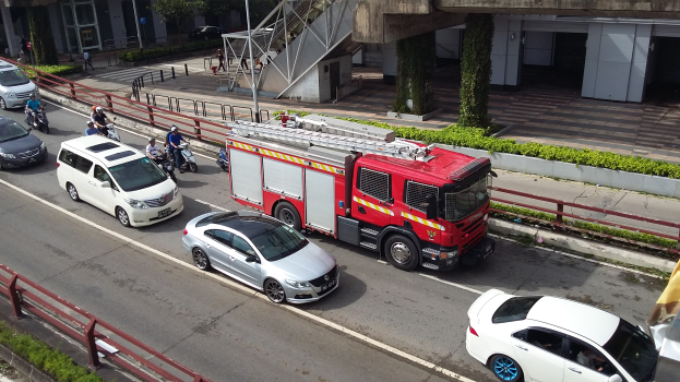 Ein Feuerwehrauto fährt eine Straße entlang neben einem Auto, während Motorräder und andere Fahrzeuge sichtbar sind. Im Hintergrund sind Gebäude, Bäume und verschiedene städtische Elemente zu sehen.