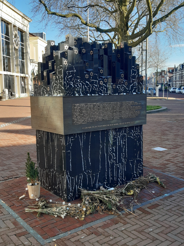 Ein Mahnmal f"{u}r Holocaust-Opfer steht auf einem Stadtplatz, mit einem Baum davor, einem Topf mit einer Pflanze und verstreuten trockenen Bl"{a}ttern, Geb"{a}uden, Fahrzeugen, Masten, B"{a}umen und dem Himmel im Hintergrund.
