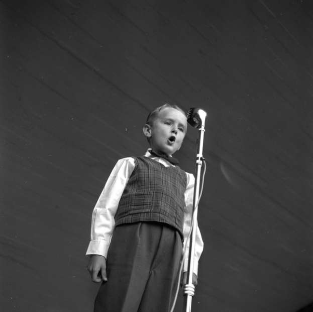 Ein junger Junge steht auf einer Bühne und singt in ein Mikrofon mit Ständer vor ihm in einem Schwarz-Weiß-Bild.