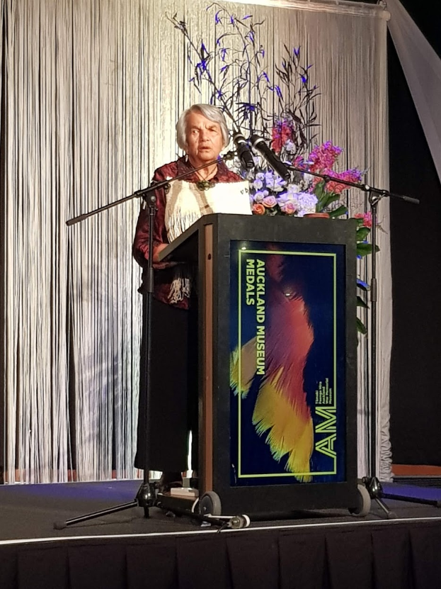 Eine Frau steht an einem Podium mit einem Mikrofon, Blumen und einer Texttafel, mit einem Vorhang und einer Wand dahinter bei der jährlichen Preisverleihung des Auckland Museums für Kunst.