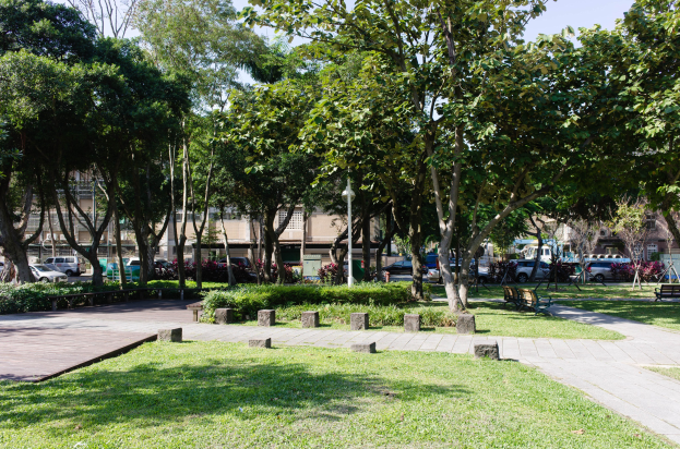 Ein Park mit grünem Gras, Bänken, Pflanzen, Bäumen, Laternenmasten und Fahrzeugen auf einer Straße, mit Gebäuden und einem klaren blauen Himmel im Hintergrund.