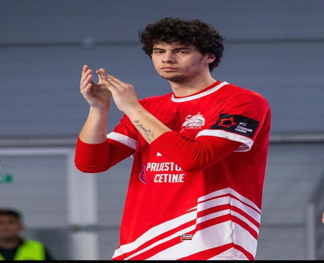 Ein Mann in einem roten und weißen Hemd klatscht in den Händen in Vordergrund, mit einer anderen Person und einer Wand im Hintergrund und Text, der unten 'Kopenhagen Futsal Club - Foto 1' liest.