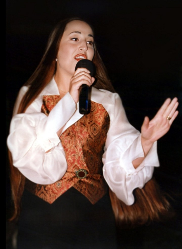 Eine Frau in einem weißen Hemd und einer braunen Weste singt selbstbewusst in ein Mikrofon und hält es fest, während ihr Haar zurückgebunden ist und ihr Blick auf das Publikum gerichtet ist.