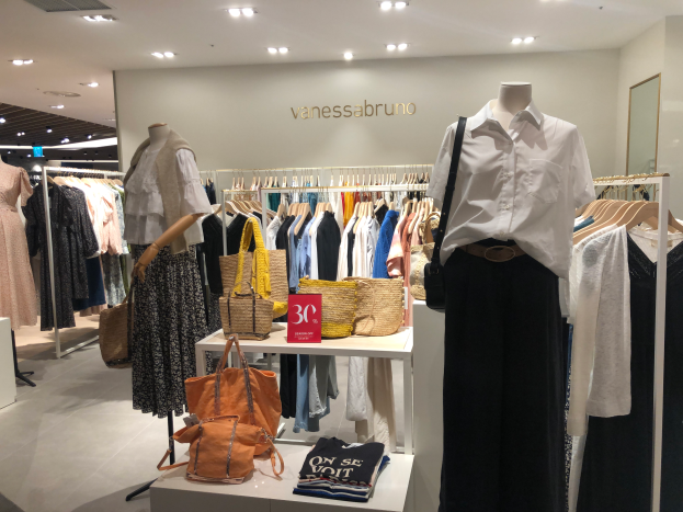 Ein Vanessa Bruno Geschäft in Singapur mit Schaufensterpuppen in Kleidern, Kleidung an Bügeln, Taschen und anderen Gegenständen auf Tischen, einem Textschild an der Wand, einem Spiegel auf der rechten Seite und Deckenbeleuchtung.