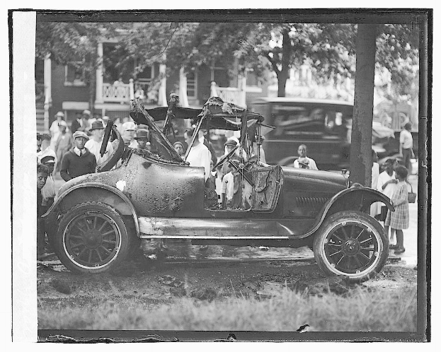 Ein Schwarz-Weiß-Foto eines feuerbeschädigten Autos mit Menschen darin, die stehen und sitzen, mit Bäumen und einem Gebäude im Hintergrund.