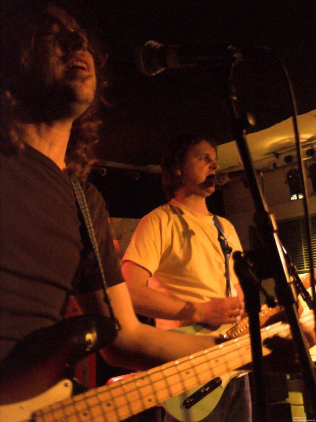Eine Person in einem weißen Hemd singt in ein Mikrofon und spielt eine grüne Gitarre, während sie neben einer anderen Person in einem schwarzen Hemd und Brille steht, die ebenfalls singt und eine schwarze Gitarre spielt, mit Scheinwerfern und Musikinstrumenten im Hintergrund.