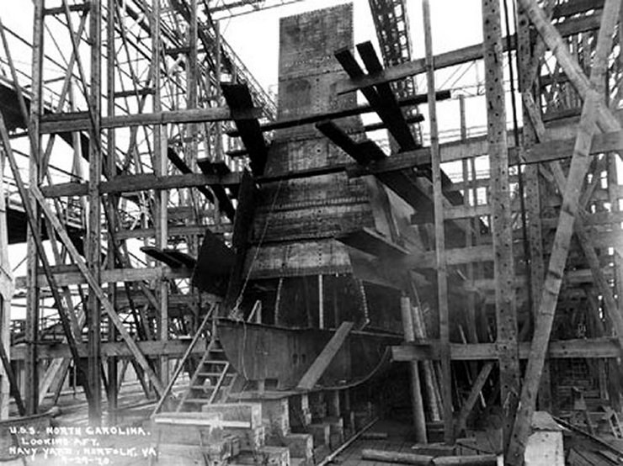 Schwarzes und weißes Foto eines Schiffes in einem Schiffbauhafen, mit Holzpfählen und Treppen im Vordergrund und Himmel im Hintergrund, mit dem Text "Marinebewaffnung" in der linken unteren Ecke.