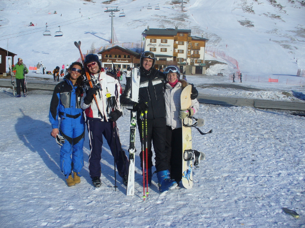 Vier Personen posieren für ein Foto mit Schlittschuhschutz und Werkzeugen, mit Gebäuden und anderen Menschen im Hintergrund, vor einer schneebedeckten Berglandschaft.