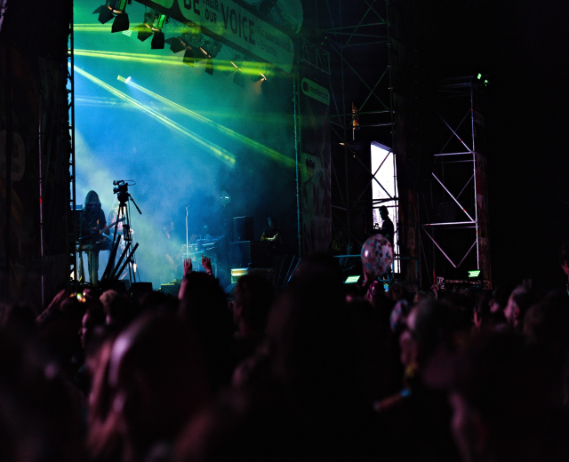 Eine Menge schaut einer Band zu, die auf einer hell erleuchteten Bühne mit Musikinstrumenten, Mikrofonen und einem Banner spielt.