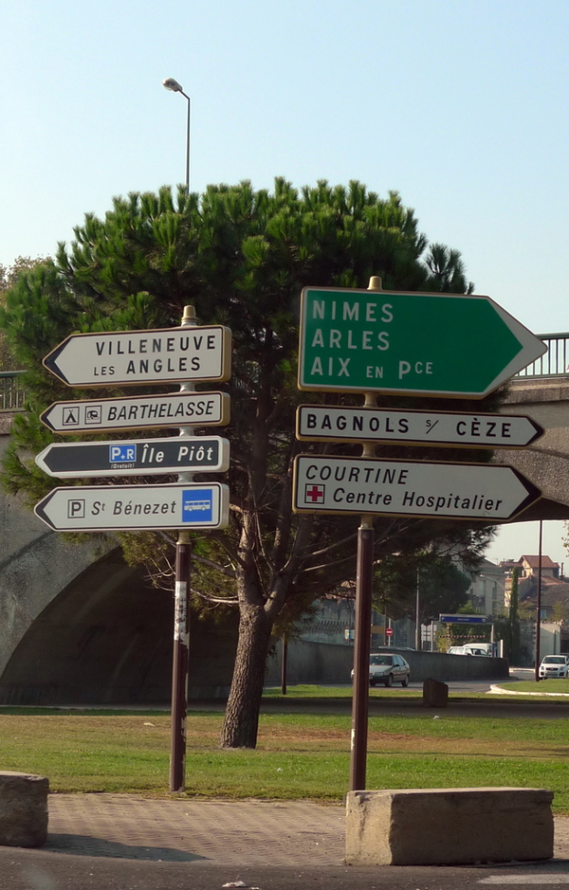 Schilder mit Text an Pfählen im Vordergrund, mit einer Brücke, Bäumen, Gebäuden, Pfählen und Fahrzeugen auf einer Straße im Hintergrund, Bänken im Vordergrund, Himmel oben und Gehweg und Gras unten.