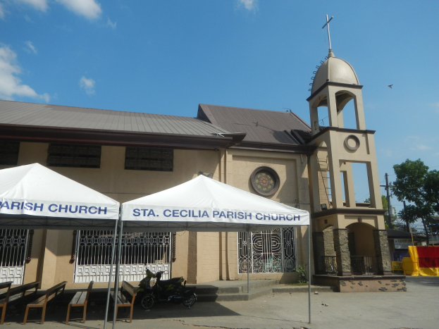 Außenansicht der St. Cecilien-Pfarrkirche in Cebu City, Philippinen, mit ihrer Fassade mit Fenstern, einer Uhr und umgeben von Bäumen, Pfählen und Drähten, mit einem Zelt und einem Motorrad im Vordergrund unter einem bewölkten Himmel.