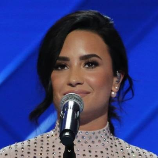 Demi Lovato in einem schwarzen Kleid mit einem eleganten Bob steht selbstbewusst vor einem Mikrofon auf der Bühne bei den Grammys, mit einem blauen Hintergrund.