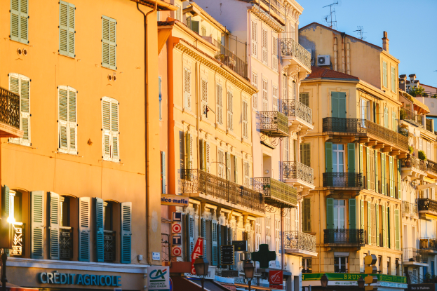 Eine Stadtstraße mit hohen Gebäuden, die Fenster, Balkone und Schilder aufweist, gesäumt von Bäumen und Straßenlaternen unter einem klaren blauen Himmel.