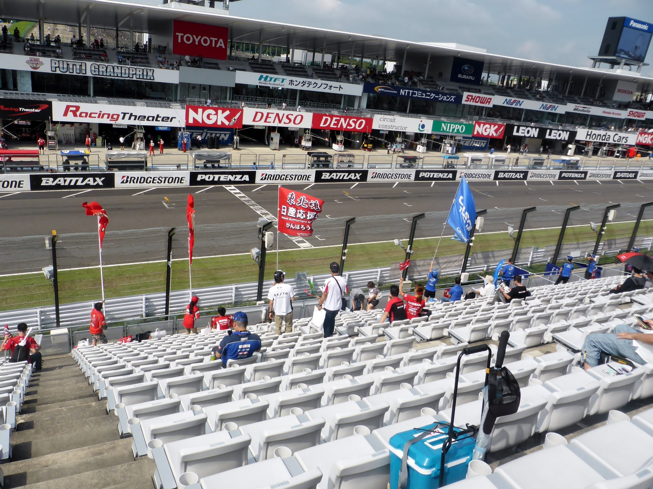 Eine Gruppe von Menschen sitzt auf Tribünen eines Rennstadions mit Fahnen, Schildern, Geländern und Stufen, mit einem bewölkten Himmel im Hintergrund.