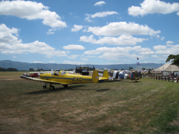 Flugzeuge, Fahrzeuge, Zelte und Fahnenmasten sind auf einer grünen Fläche mit Bäumen und Hügeln im Hintergrund unter einem klaren Himmel verstreut.