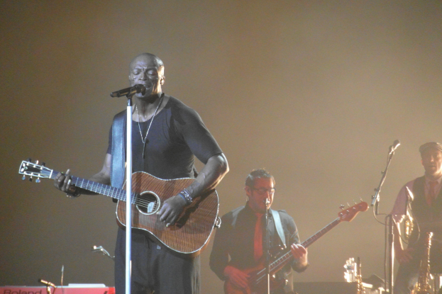Ein Mann auf der Bühne, der Gitarre spielt und in ein Mikrofon singt, begleitet von Musikern, die Saxofon und andere Instrumente spielen.