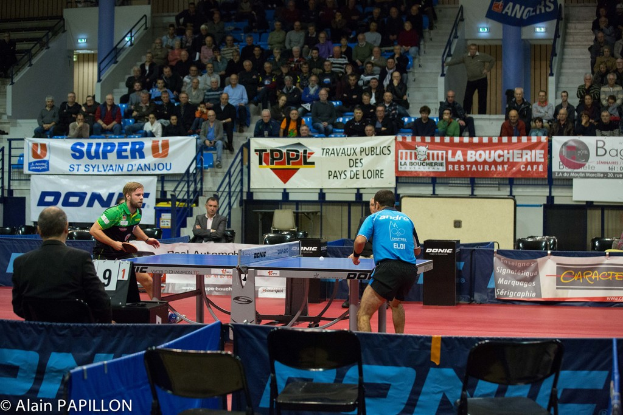 Zwei Männer spielen Tischtennis drinnen mit Zuschauern, die stehen und sitzen, leere Stühle und Werbeplakate, mit Text in der unteren linken Ecke.