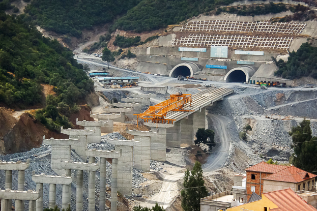 Eine Baustelle mit einem großen Tunnel, umgeben von Bäumen, Gebäuden, Fahrzeugen, Pfeilern, Felsen und anderen Gegenständen, mit einer Straße am unteren Bildrand.