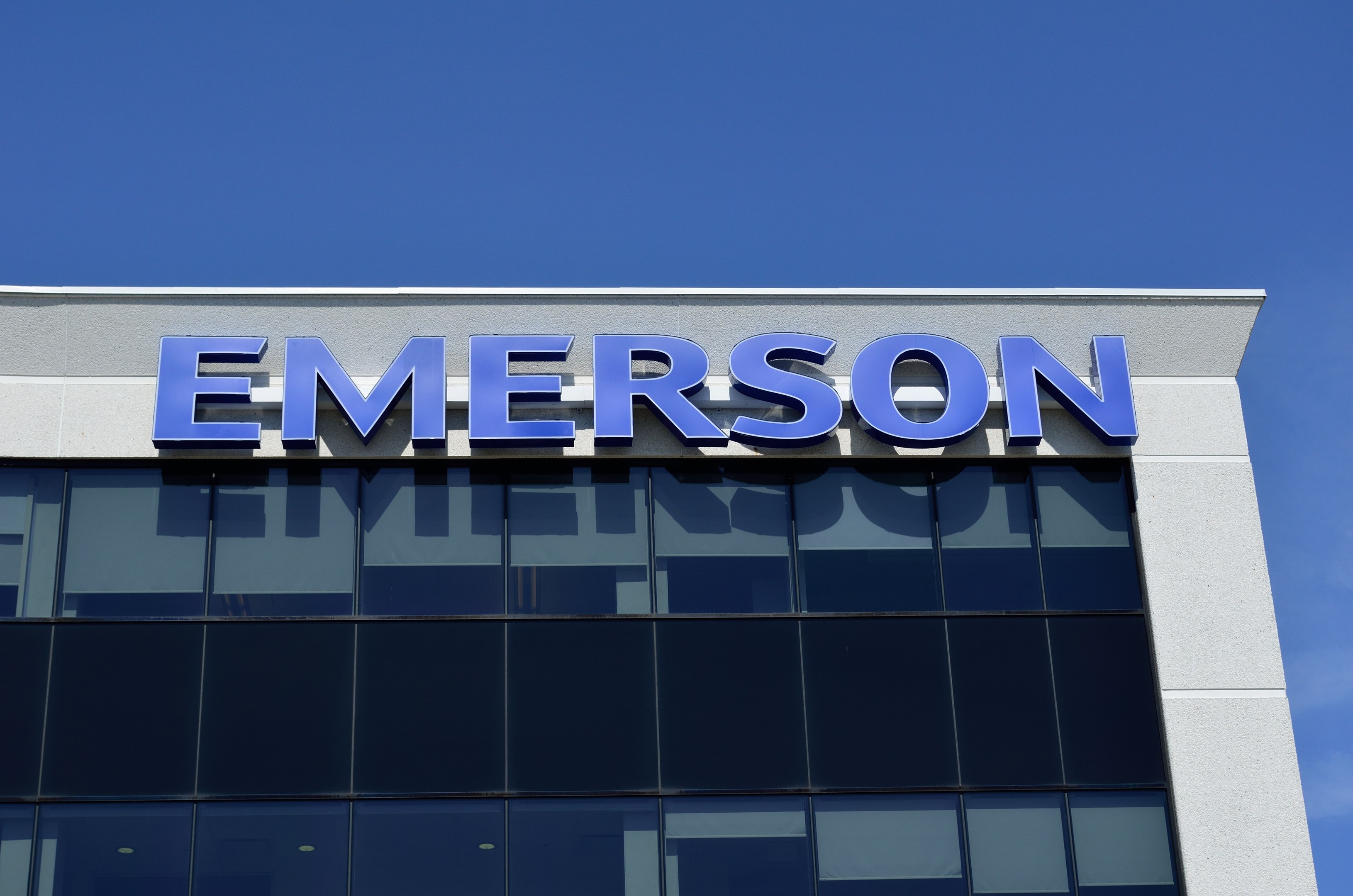 Das Emerson-Logo an der Seite eines Gebäudes mit dem Text "Emerson investiert 1,5 Milliarden USD in den USA" vor einem klaren blauen Himmel.
