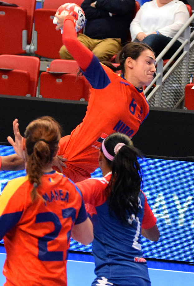 Eine Gruppe von Frauen, die in einem Stadion Handball spielt, wobei eine den Ball hält und Zuschauer im Hintergrund sitzen.