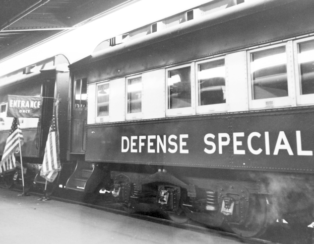 Schwarzes und weißes Foto eines Zuges mit der Aufschrift "Defense Special" auf einem Bahngleis, mit zwei Fahnen links und einer Decke darüber.