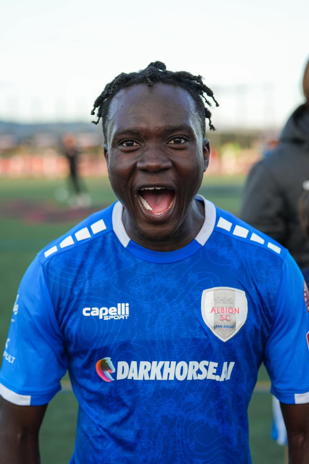 Ein Mann in einem blauen T-Shirt steht auf einem Fußballfeld und hat den Mund offen, wahrscheinlich feiert er ein Tor von Albion FC, mit ein paar unscharfen Menschen im Hintergrund.