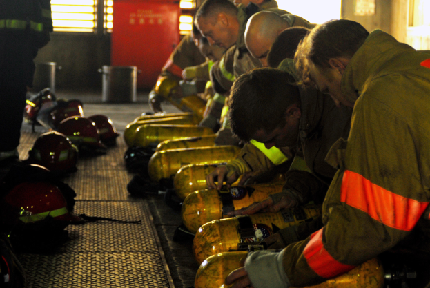 Feuerwehrleute arbeiten an einem Feuerwehrauto mit Ausrüstung und Helmen, die herumliegen, in einem Raum mit Fenstern, einer Tür und einem Müllcontainer.