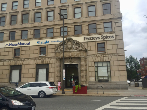 Großes Gebäude mit parkenden Autos davor, Straßenlaternen, Fahrzeugen, Fußgängern, Pflanzen, Bäumen und bewölktem Himmel, identifiziert als Hauptsitz der Massachusetts Mutual Financial Group.