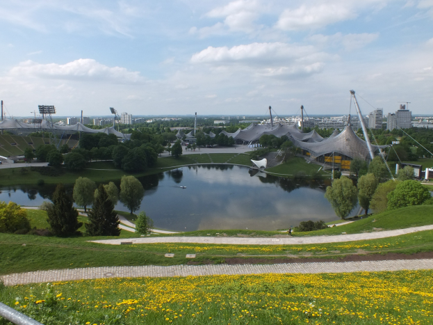 Ansicht des Olympiastadions von einem Hügel aus, mit einem Teich im Vordergrund, umgeben von Grünflächen und gelben Blumen, Gebäuden und Pfählen im Hintergrund unter einem klaren blauen Himmel.