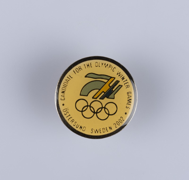 Ein rundes Abzeichen mit schwarzem Hintergrund und goldfarbenem Olympia-Winterspiellogo, mit der Beschriftung "Olympische Winterspiele" in fetter weißer Schrift auf einer weißen Oberfläche.