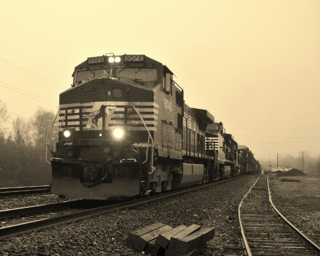 Ein Zug auf Eisenbahnschienen mit Autos, Strommasten, Böumen und Himmel im Hintergrund.