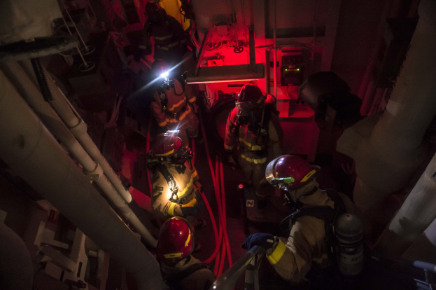 Feuerwehrleute in Helmen und Sauerstoffflaschen stehen in einem dunklen Raum mit Rohren auf beiden Seiten, bereit für einen Rettungseinsatz in der Nähe von Maschinen im Hintergrund.