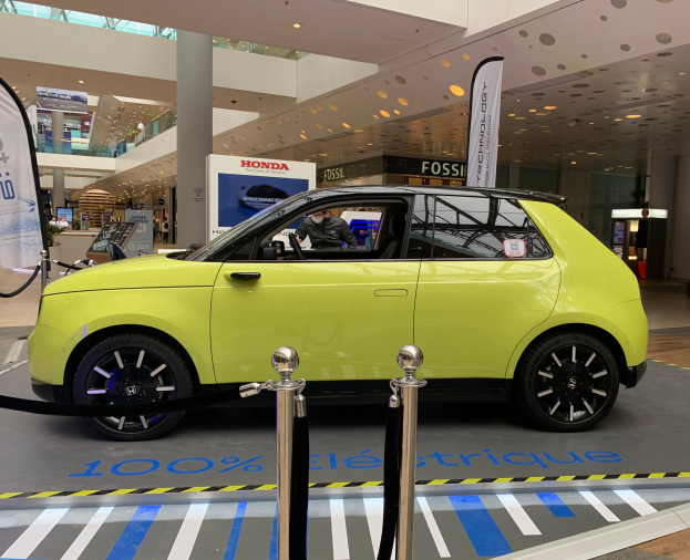 Gelber wasserstoffbetriebener Elektroauto vor einem Gebäude mit Absperrpfosten, Bannern, Menschen, Schildern, einer Topfpflanze, Deckenleuchten und einer Decke mit Lüftern.