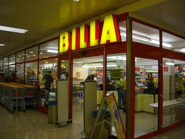Ein Markt mit einem Eingang in der Mitte, Mülltonnen daneben, einer Reihe von Einkaufswagen auf der linken Seite, Deckenbeleuchtung und einigen Menschen, die Produkte betrachten.