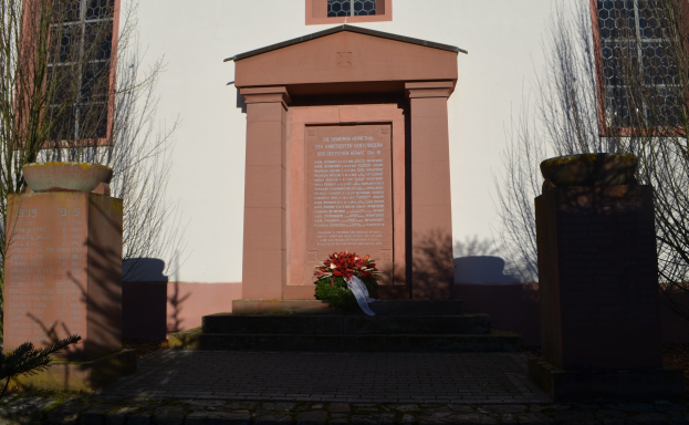 Ein Gebäude mit Fenstern, ein Denkmal mit Text, ein Blumenstrauß auf dem Boden, Topfpflanzen, eine Baumgruppe, ein bewölkter Himmel und ein Grab mit einem Kranz davor.